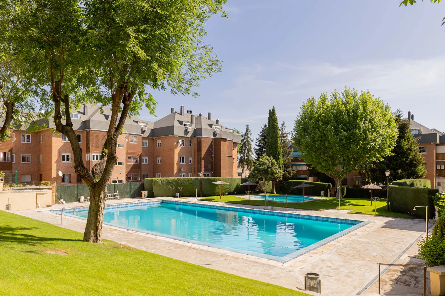 Outdoor pool with turquoise water, surrounded by green lawn and trees, in front of a brick apartment building.