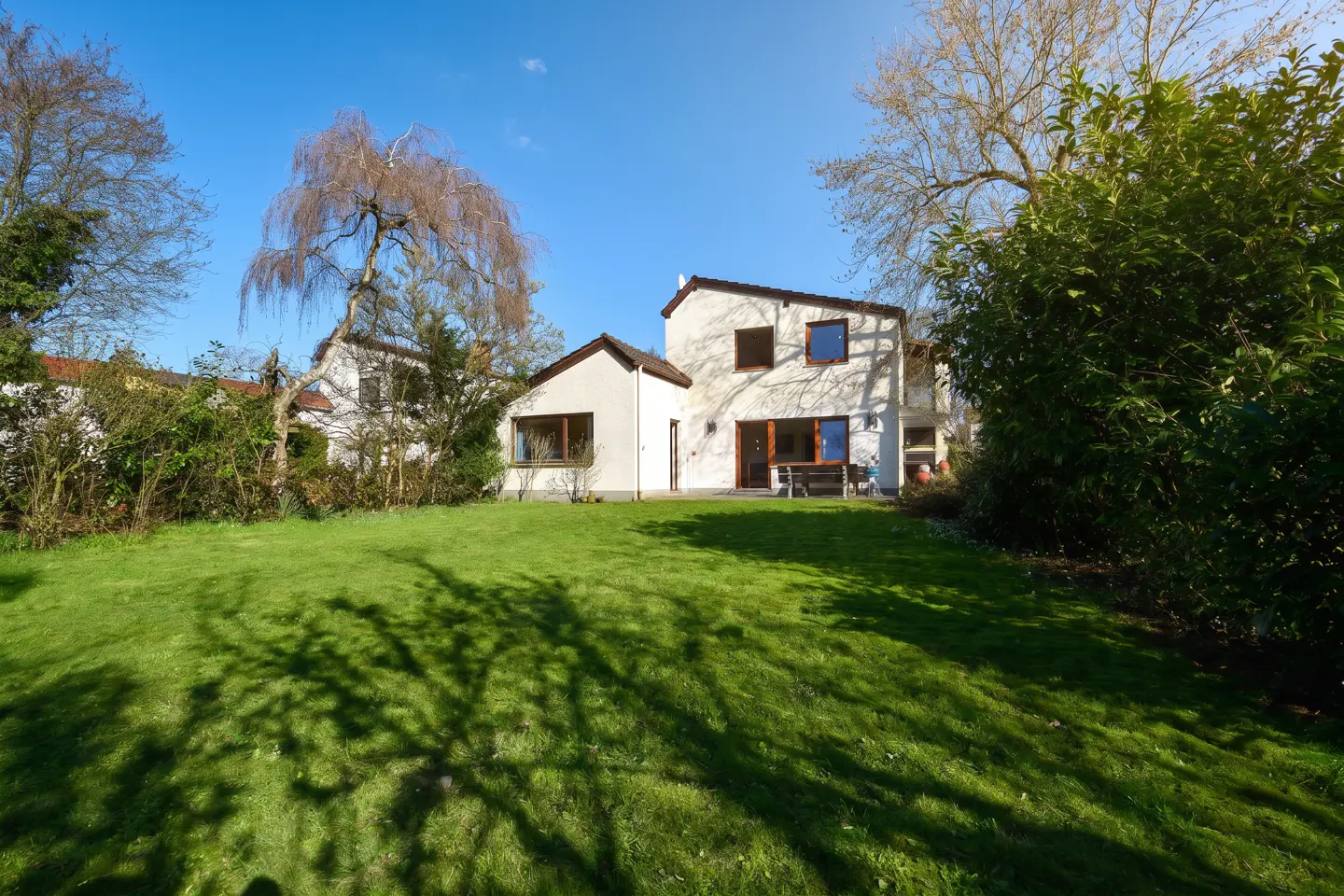 Back view of a white two-story house with a large green lawn, surrounded by trees under a clear blue sky.