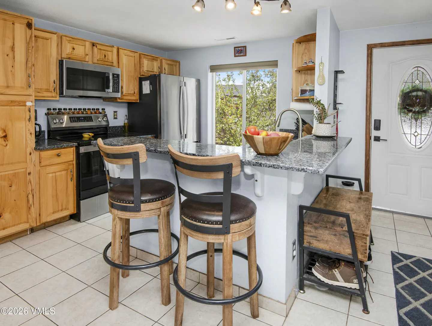 A kitchen with light wood cabinets, stainless steel appliances, and a granite countertop island with two bar stools.