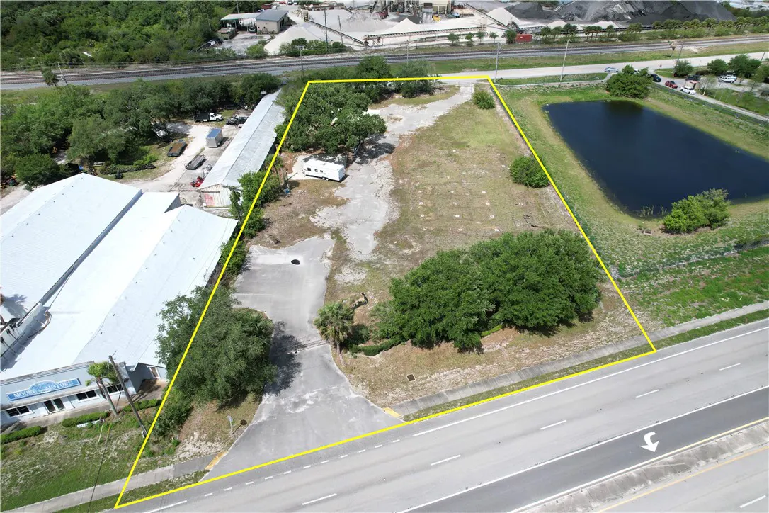 Aerial view of a vacant lot outlined in yellow, adjacent to a road, pond, and industrial buildings. Trees and some vegetation are visible on the lot.