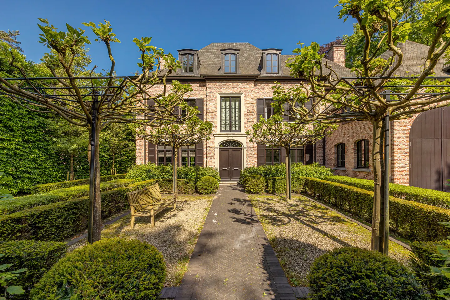 Brick house with dark shutters and a dark front door, framed by manicured trees and hedges. A stone path leads to the entrance.