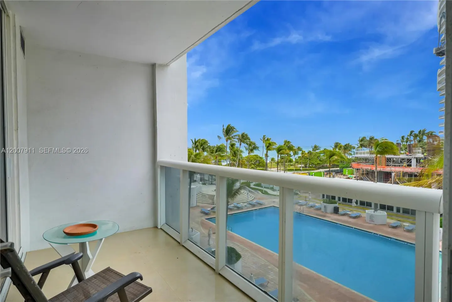 Balcony view with chairs, table, and pool. Palm trees and blue sky visible in the background.