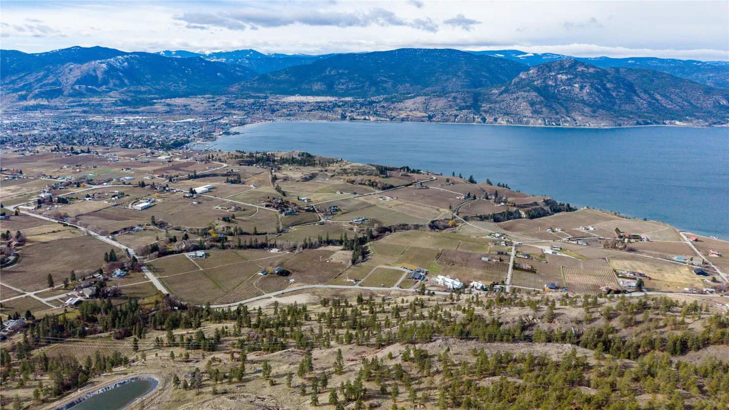 Aerial view of Okanagan Valley with lake, vineyards, and mountains in the background. Houses are scattered throughout the landscape.