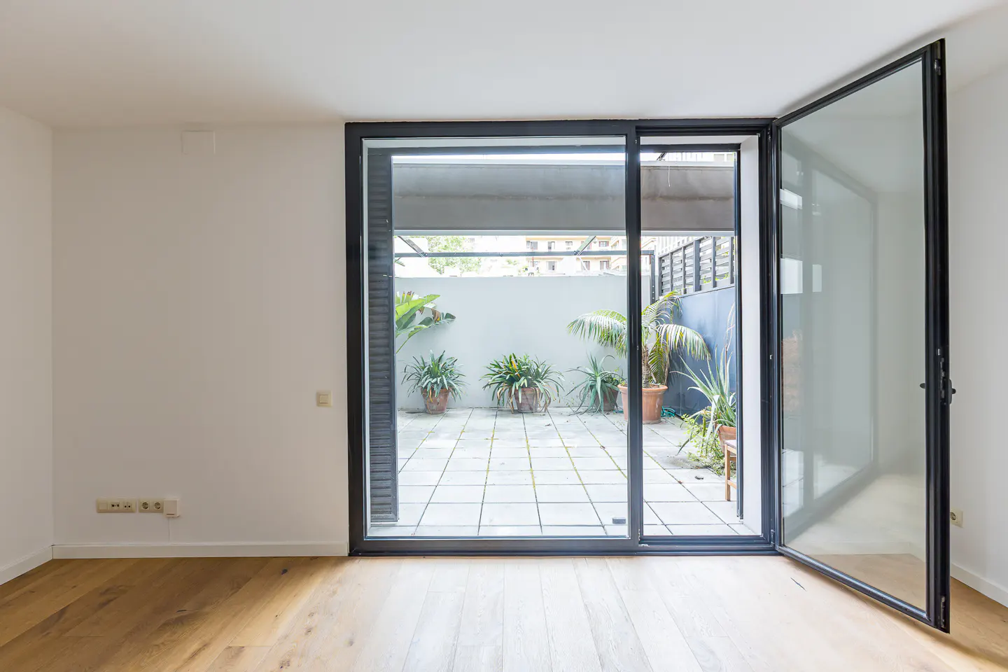 Modern room with light wood floors and large glass doors opening to a tiled patio with potted plants and a grey wall.