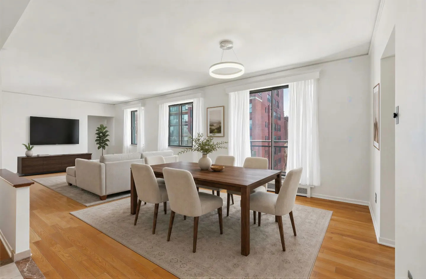 Bright, open-concept living and dining area with hardwood floors, white walls, and large windows. A wood table with six chairs sits on a neutral rug.