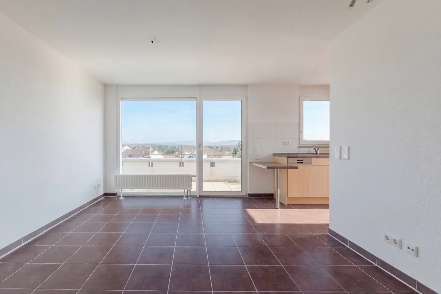 Bright, empty apartment with brown tile floor, white walls, and a view from the balcony. A small kitchen area is visible.