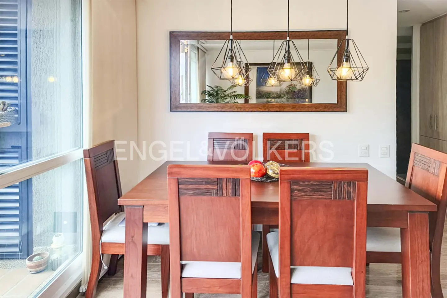 A dining room features a wooden table and chairs, with modern pendant lights above. A mirror reflects artwork and natural light from a nearby window.