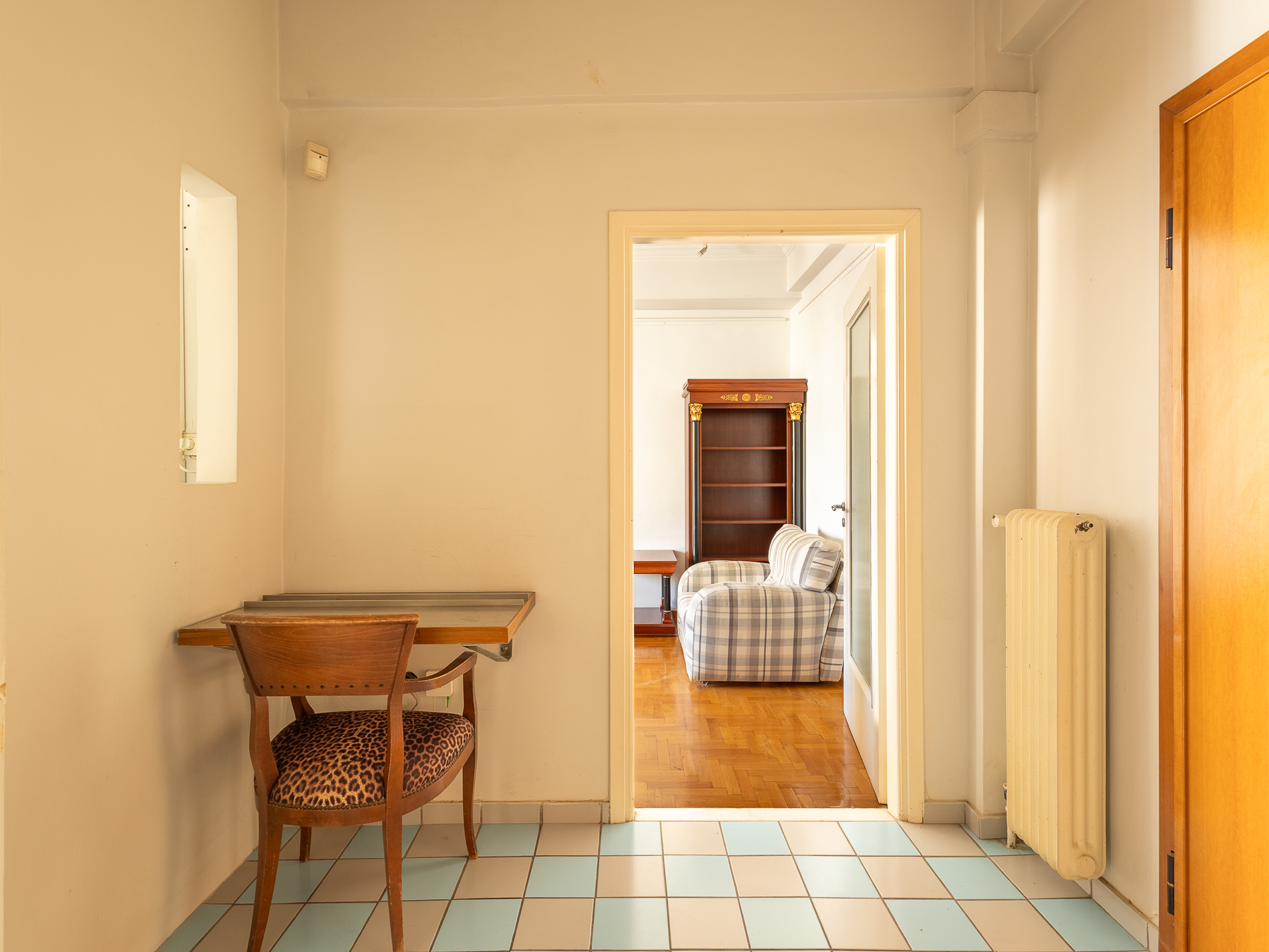 Small hallway with a wall-mounted desk, leopard-print chair, blue and white tiled floor, and a view into a living room. [AI-generated text]