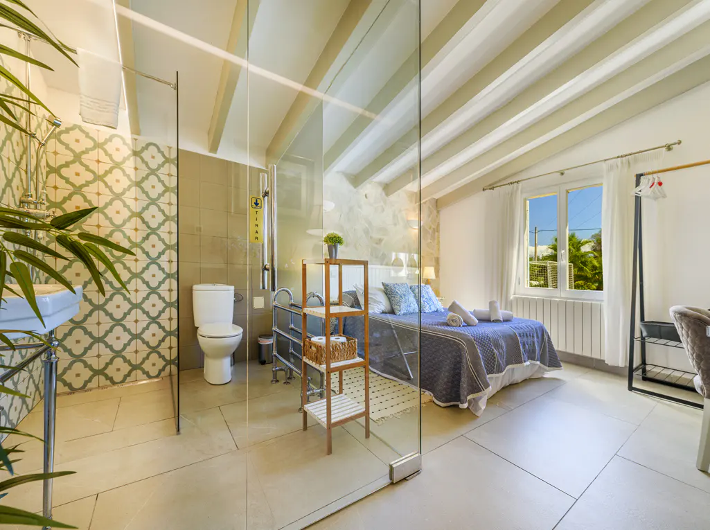 Bedroom with ensuite bathroom separated by glass. Blue quilt on bed, white walls, and patterned bathroom tiles.