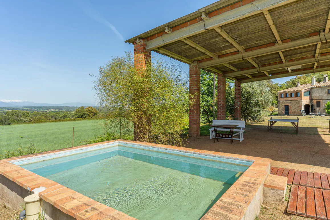 Outdoor pool with brick surround, covered patio with seating, and green field in the background.