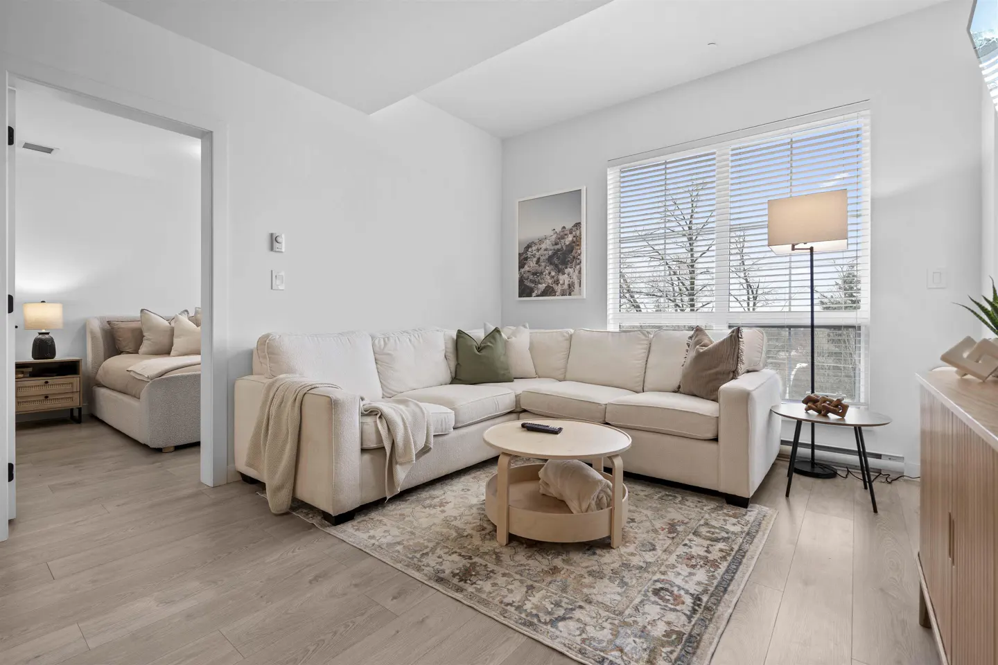 Modern living room with a cream sectional sofa, round wooden coffee table, patterned rug, and a view into a bedroom.