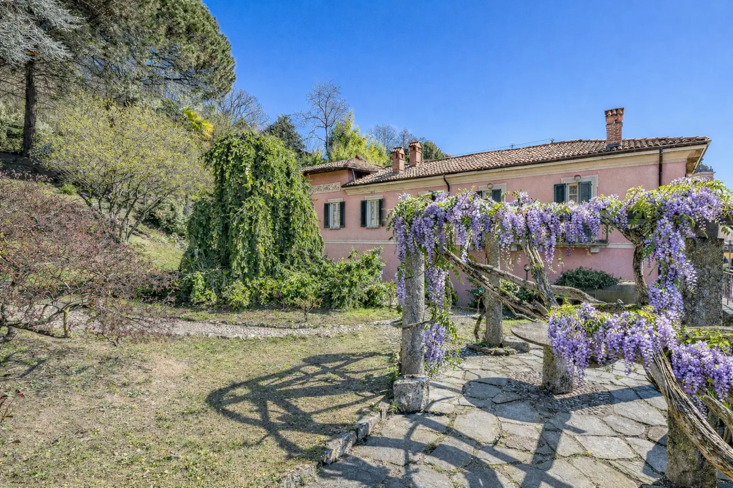 Exterior view of a pink house with green shutters, stone patio, and wisteria-covered pergola.