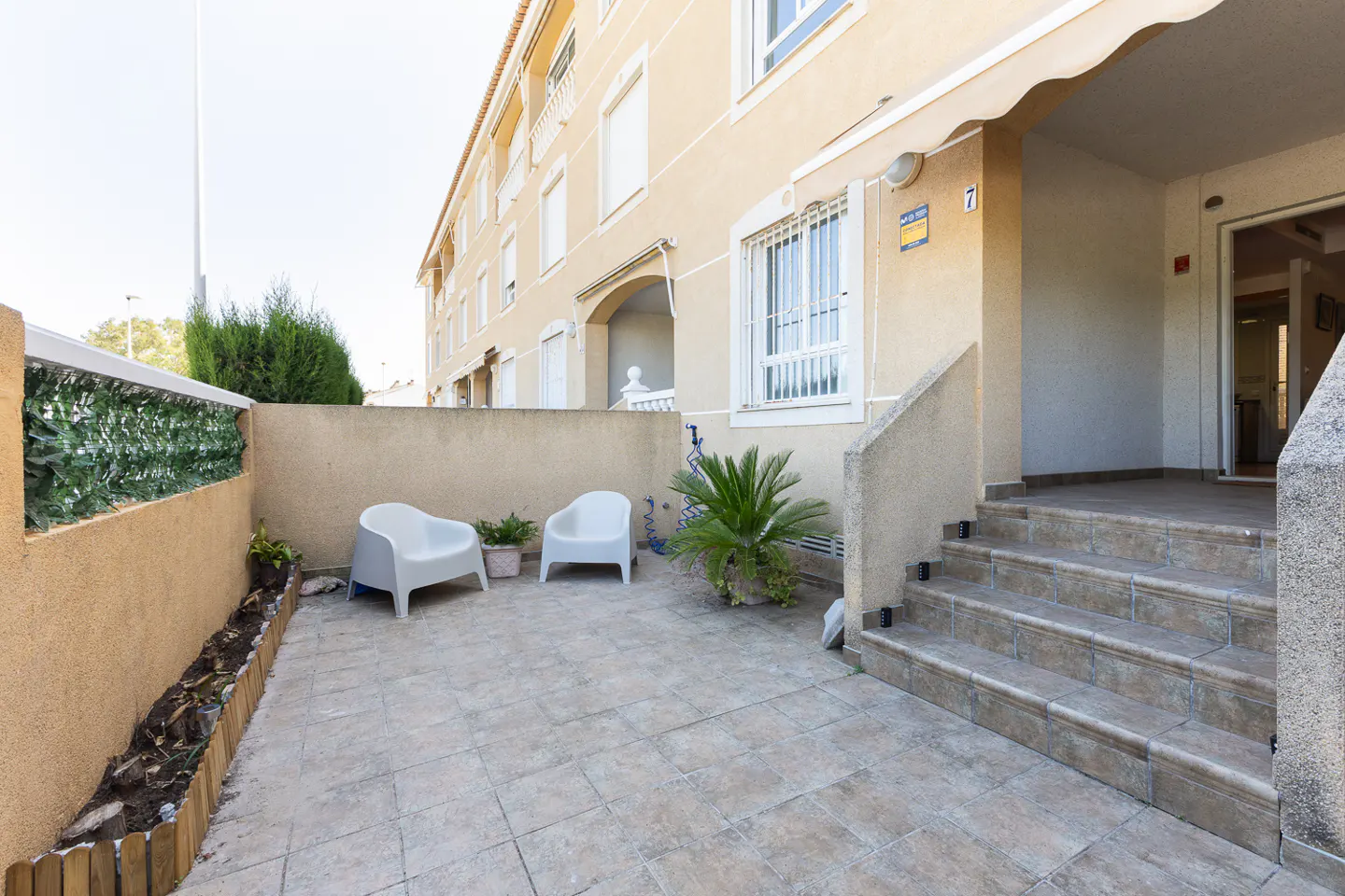 A tiled patio with two white chairs, a small potted plant, and stone steps leading to the entrance of a beige building.