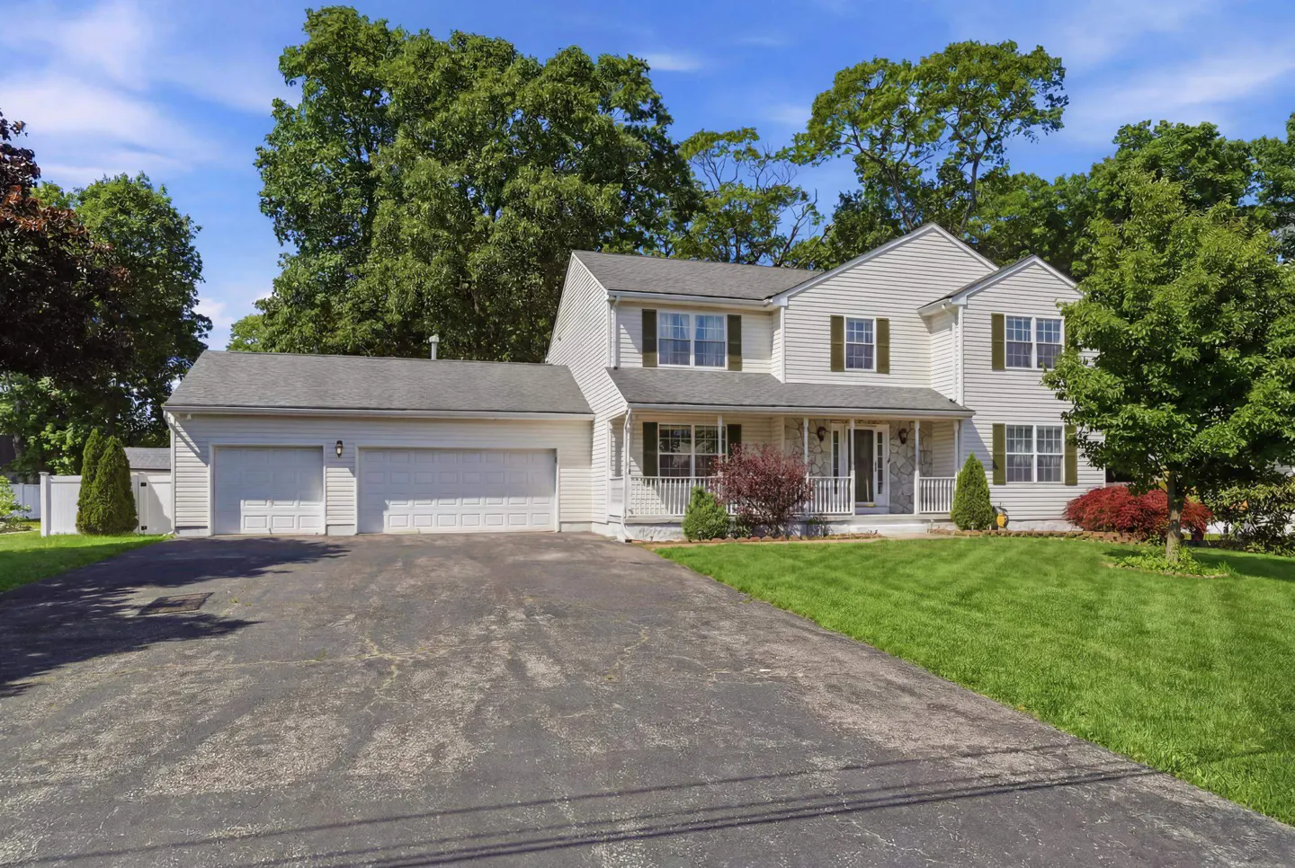Two-story beige colonial home with a three-car garage, front porch, and large green lawn under a blue sky.