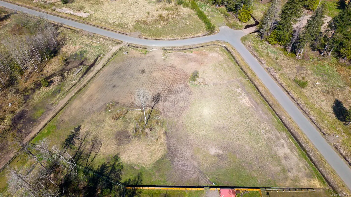 Aerial view of a large, vacant, triangular lot bordered by roads and trees. The lot is mostly brown with patches of green grass.