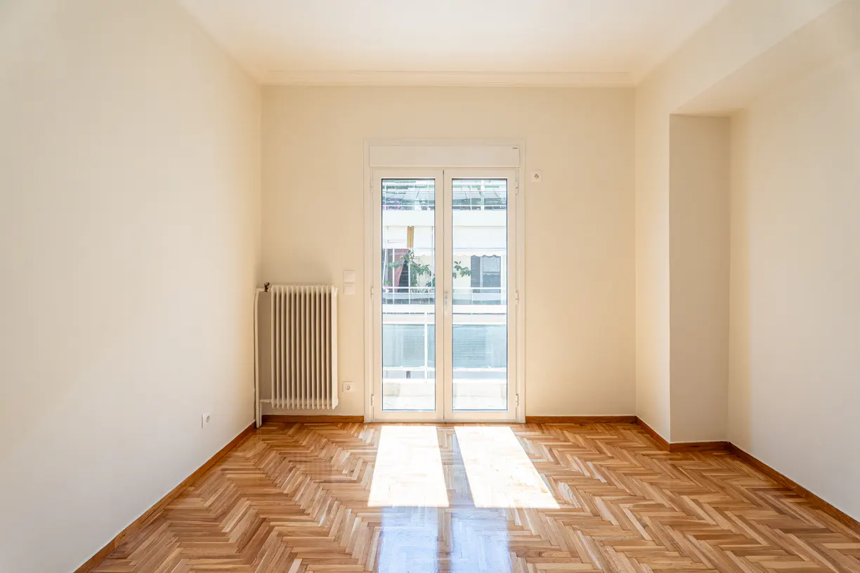 Bright, empty room with herringbone wood floors, white walls, and a radiator. Double doors lead to a balcony.