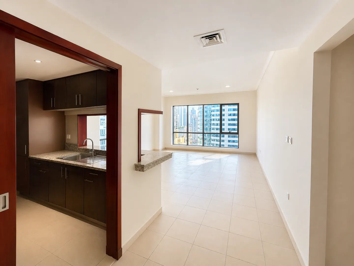 A bright, empty apartment with a kitchen view. Dark wood cabinets, granite counters, and a large window with a city view.
