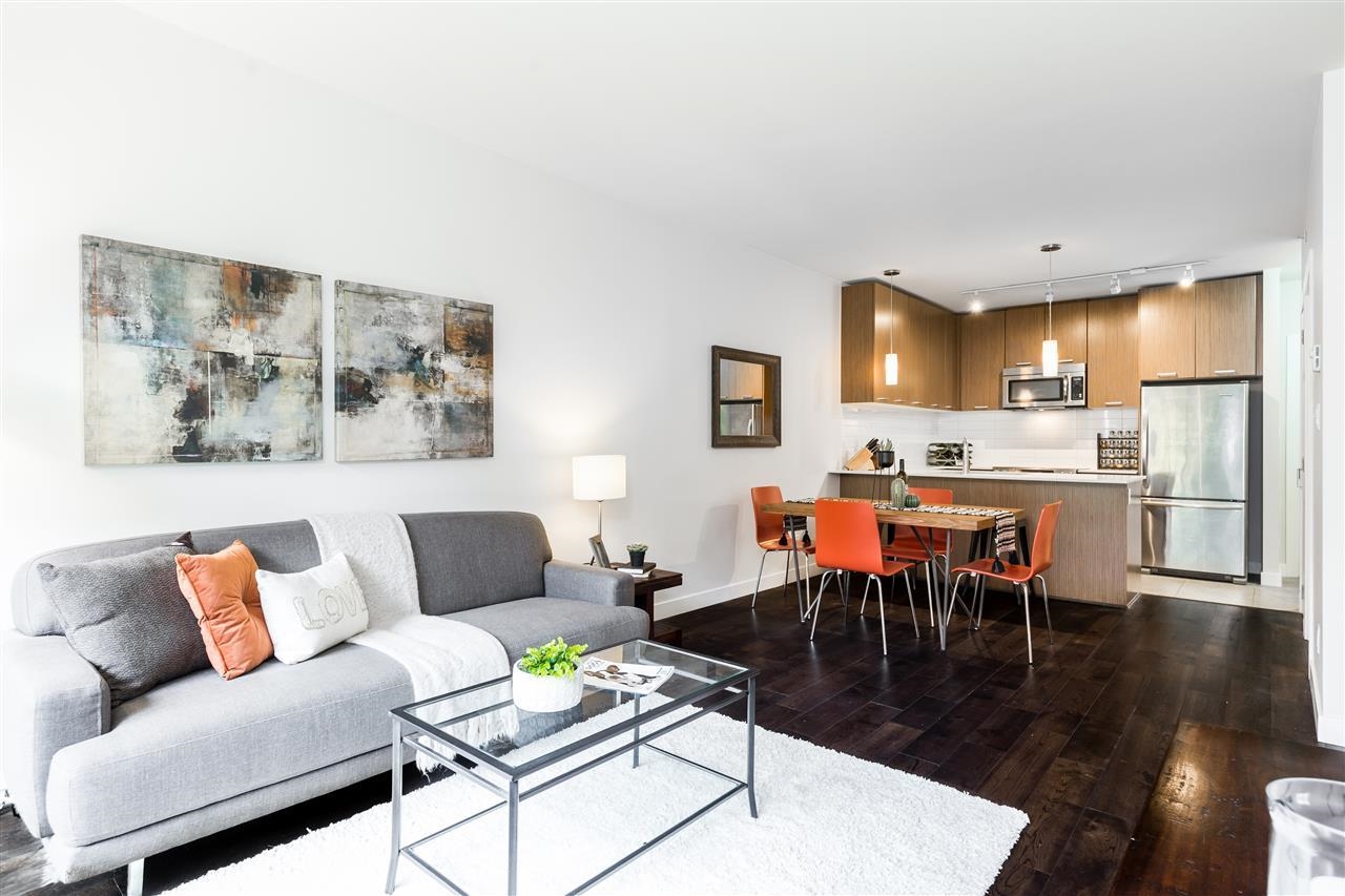 Modern open-concept living room with a grey sofa, glass coffee table, and dining area with orange chairs and wood cabinets.