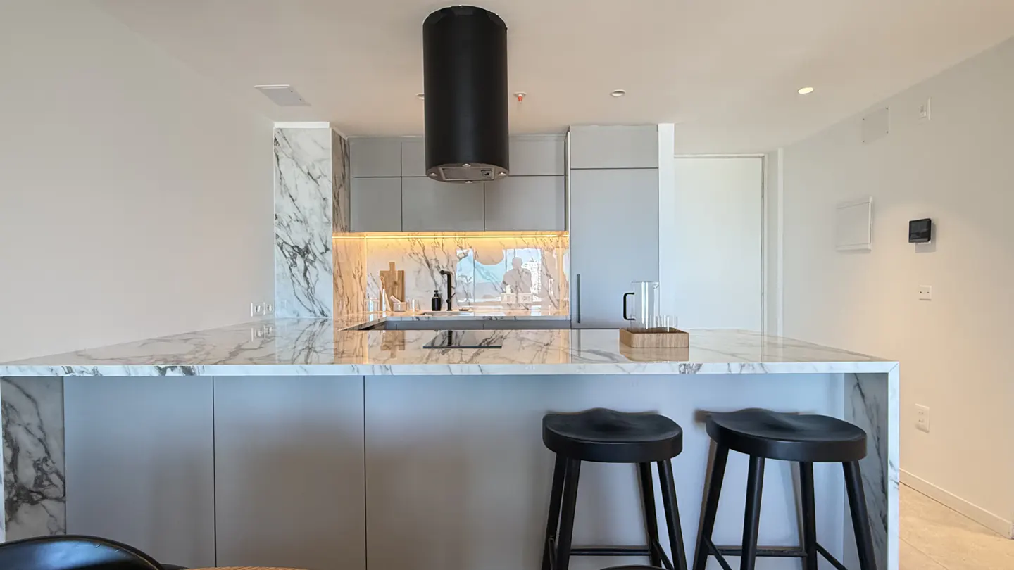 Modern kitchen with marble countertops, gray cabinets, and black stools. A black cylindrical range hood hangs above the island.