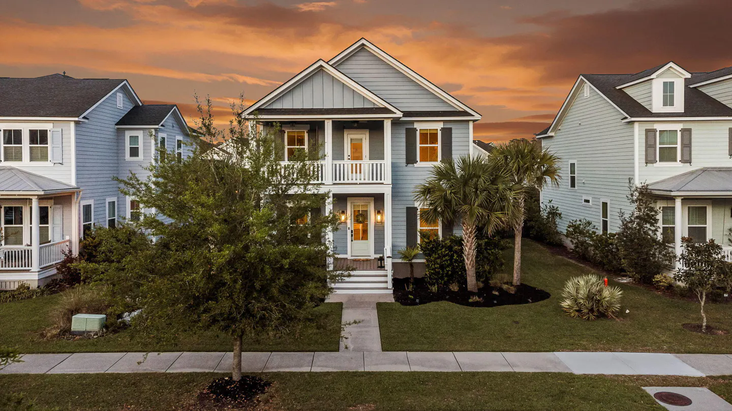 Two-story light blue house with white trim, black shutters, and a small porch. Palm trees and green lawn in front. Sunset sky.