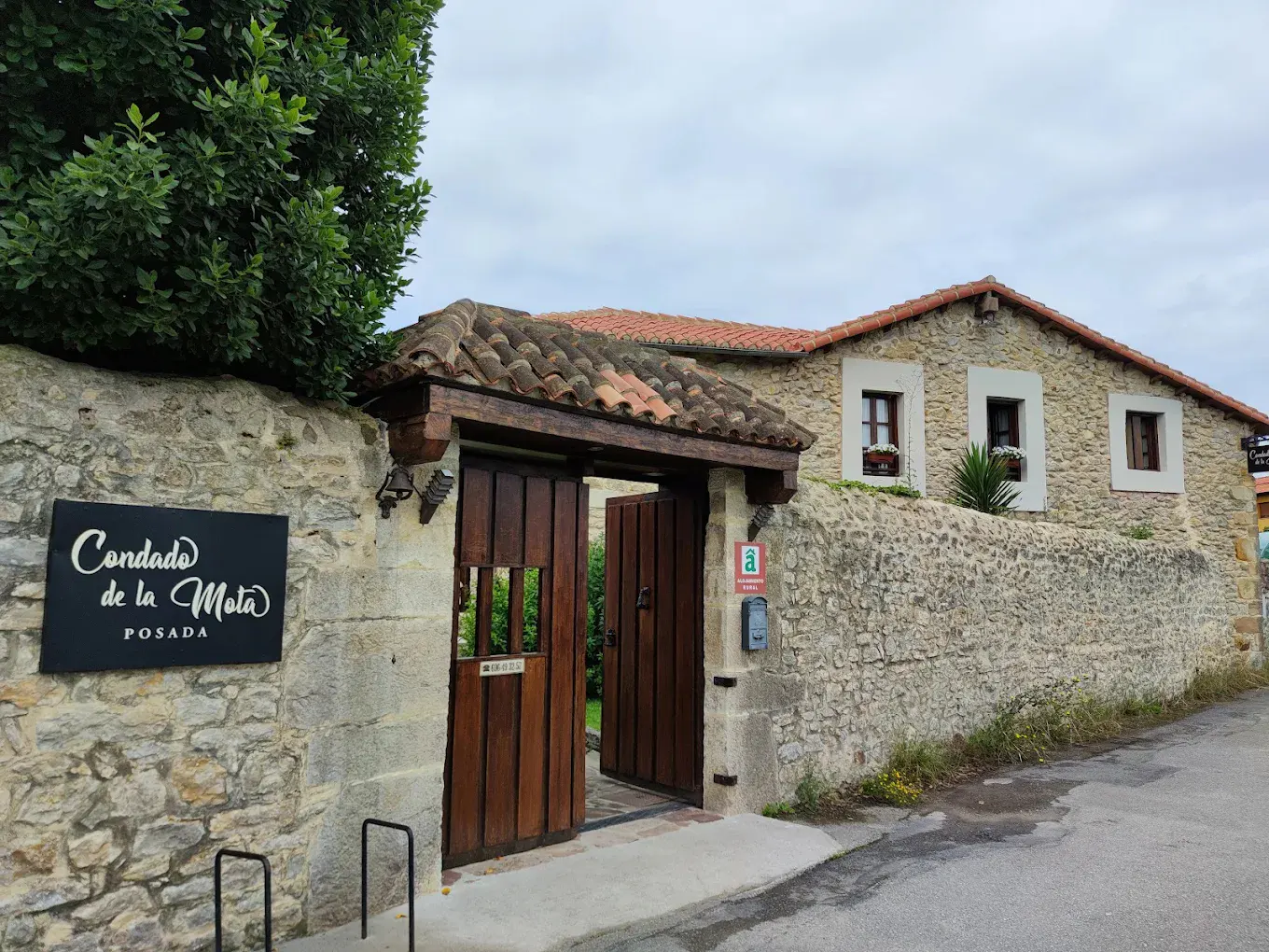 Stone building with red tile roof, "Condado de la Mota" sign, and open wooden gate in a stone wall.