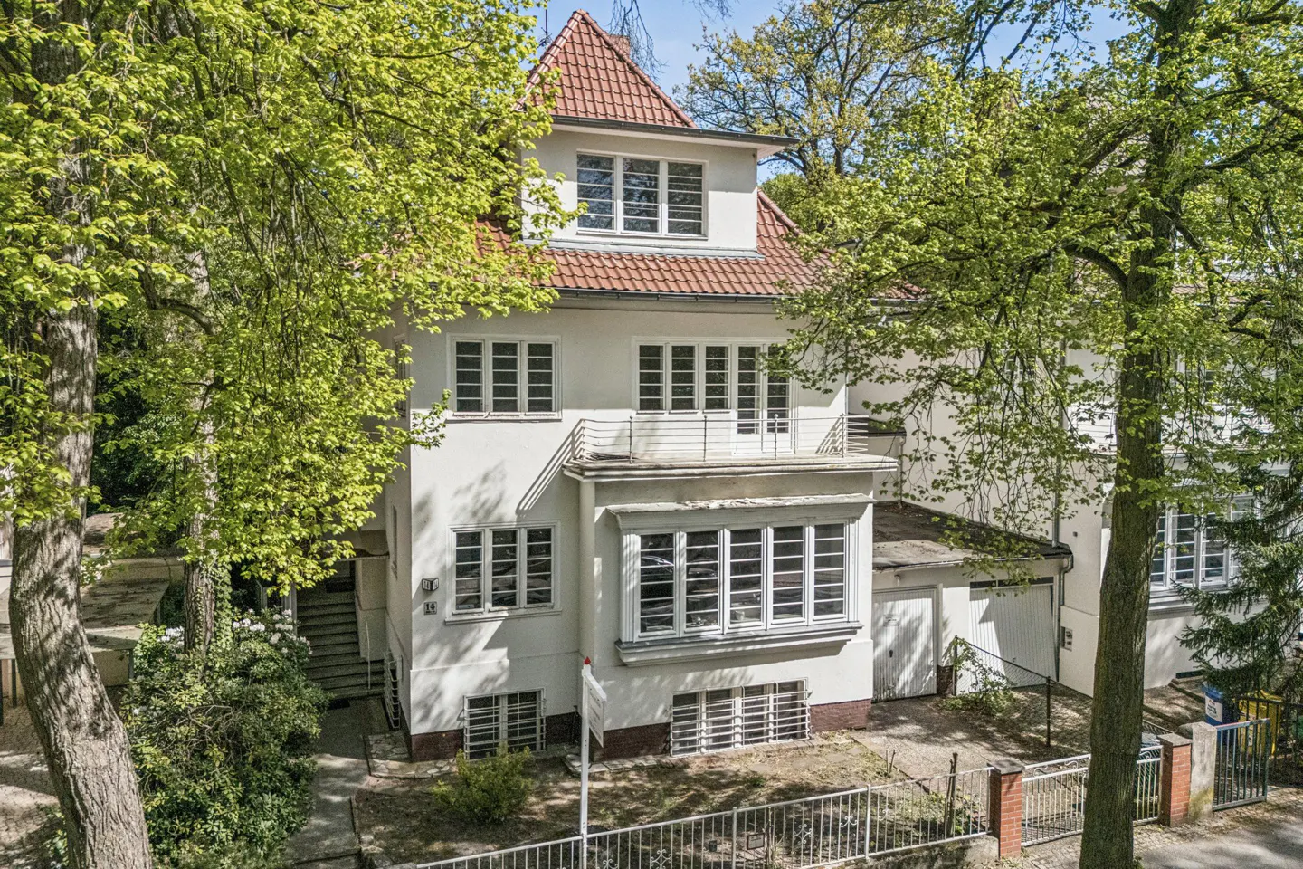 A three-story white house with a red tile roof is surrounded by green trees. The house has many windows and a balcony. A metal fence surrounds the property.