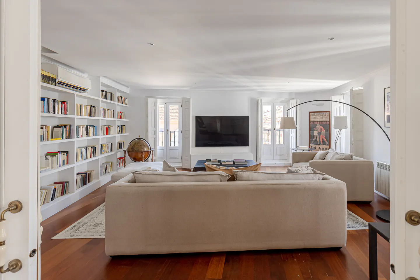 Bright living room with hardwood floors, white walls, and a large bookshelf filled with books. Two sofas face a TV, with French doors in the background.