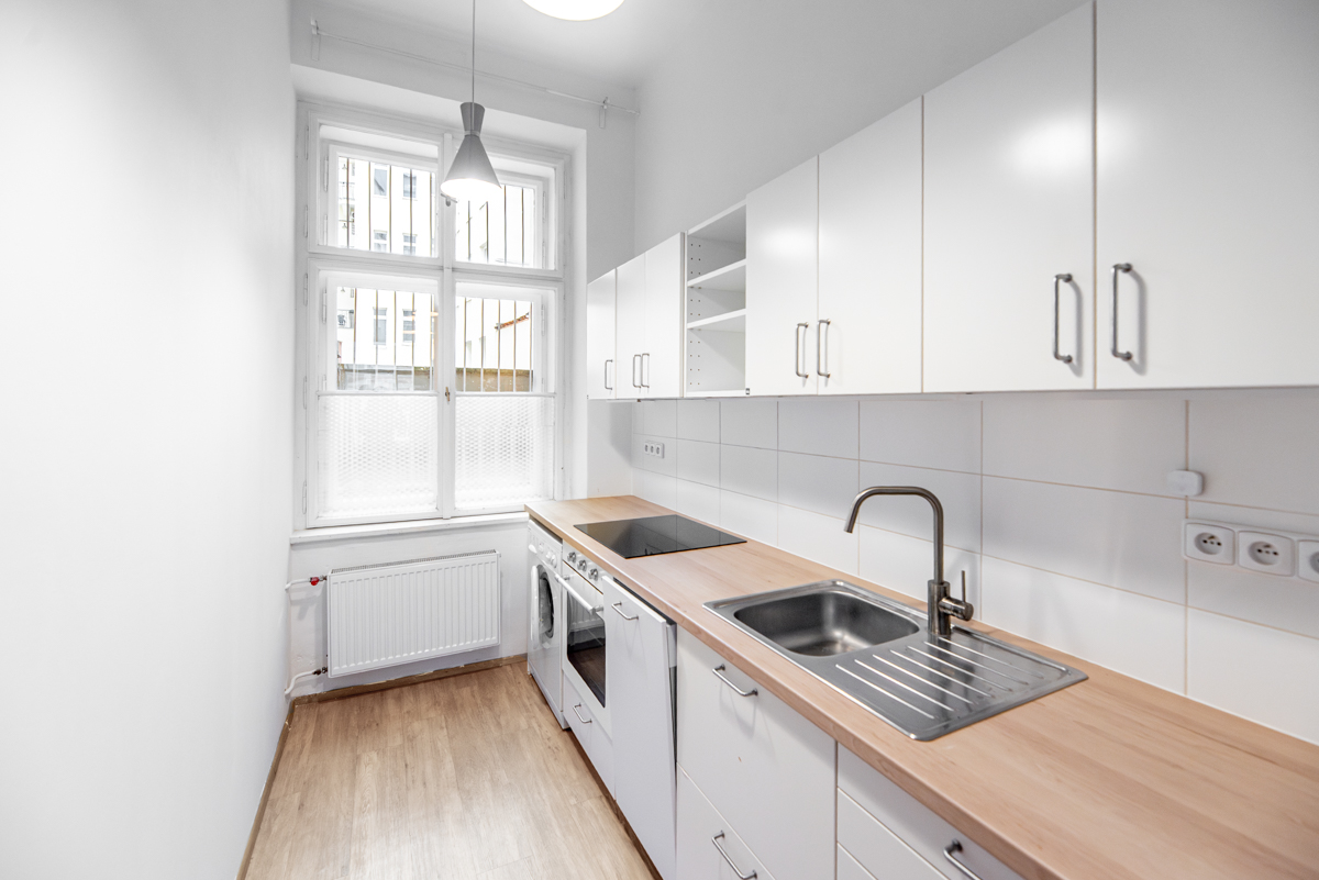 Bright, narrow kitchen with white cabinets, wood countertop, and stainless steel sink. A window with bars is on the left.