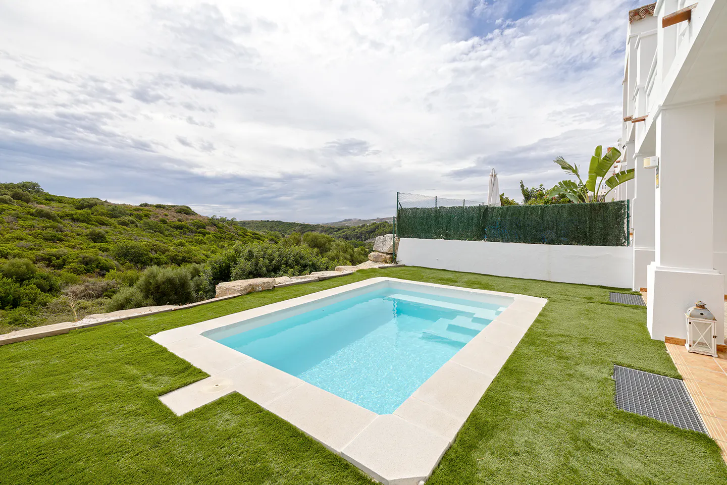 A rectangular swimming pool set in a green lawn with a scenic view of rolling hills under a cloudy sky.