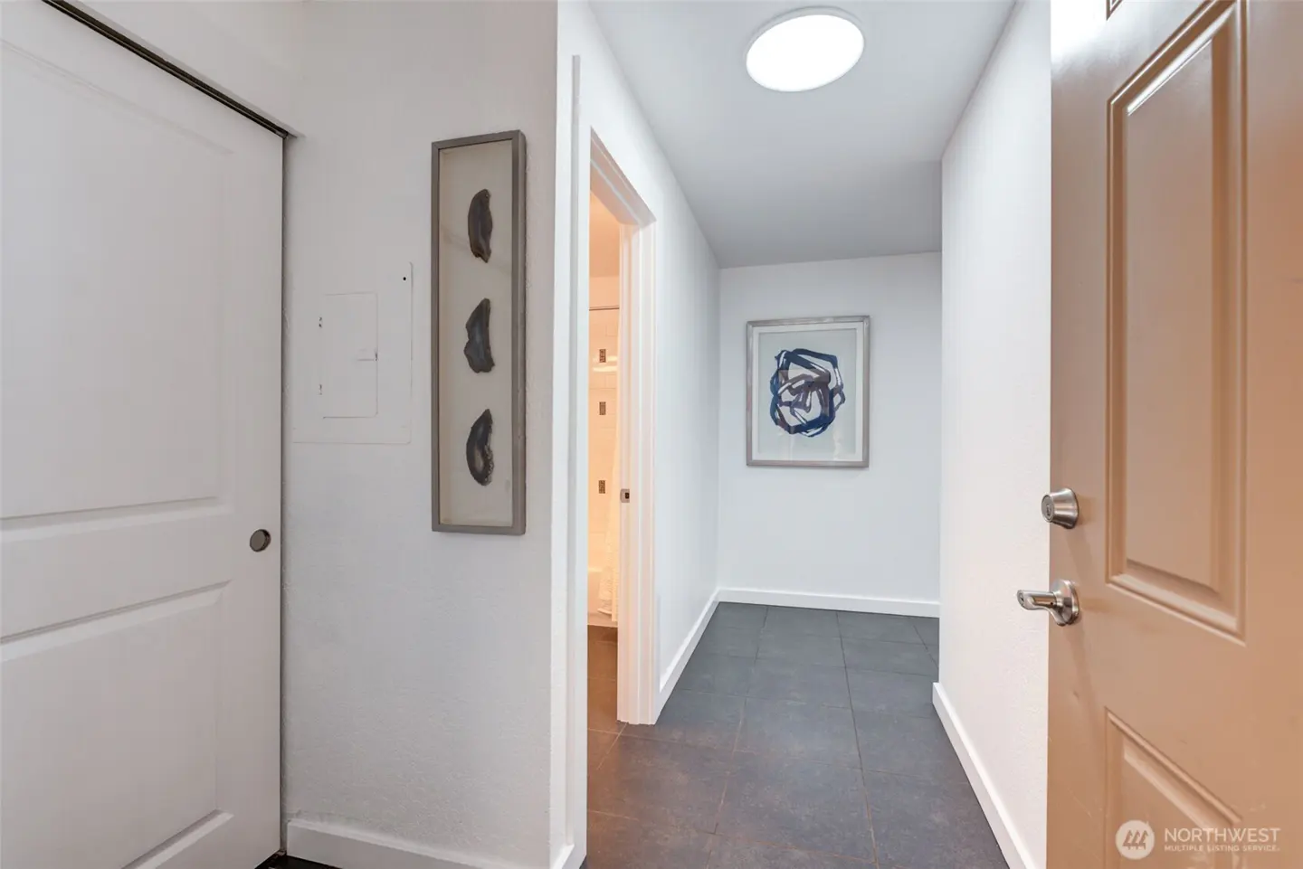 Bright hallway with white walls, gray tile floor, and a skylight. Artwork hangs on the walls, and doors lead to other rooms.