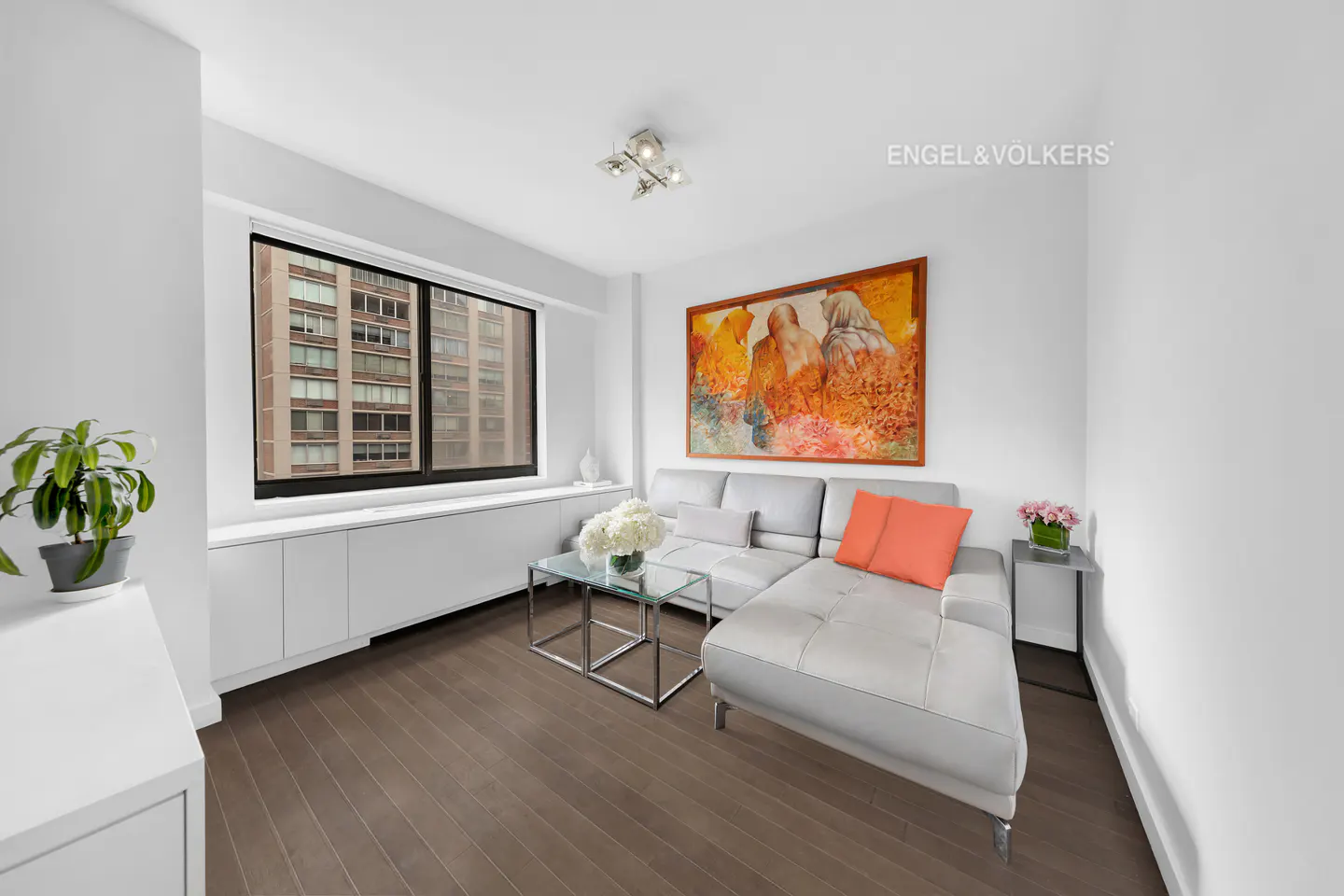 Bright living room with hardwood floors, white walls, and a gray sectional sofa with an orange pillow. A large window overlooks a city building.