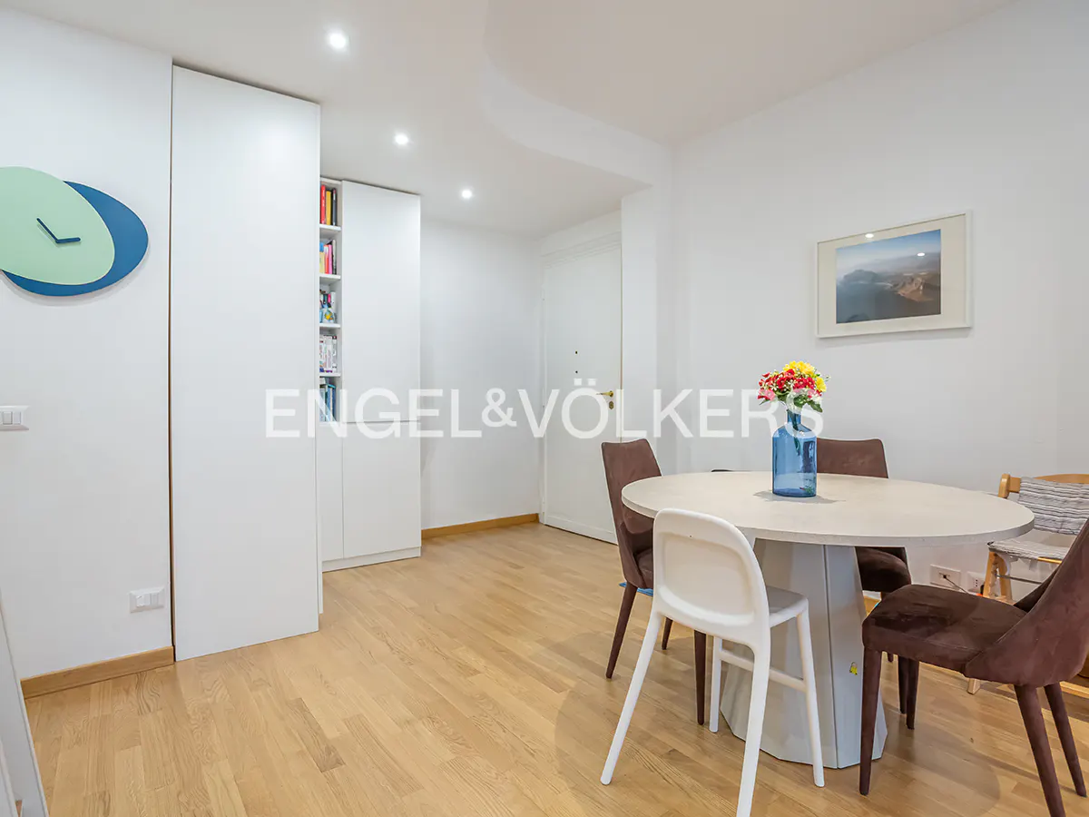 Bright dining area with a round table, brown and white chairs, and a blue vase with flowers. White walls and wooden floors. Engel & Völkers logo.