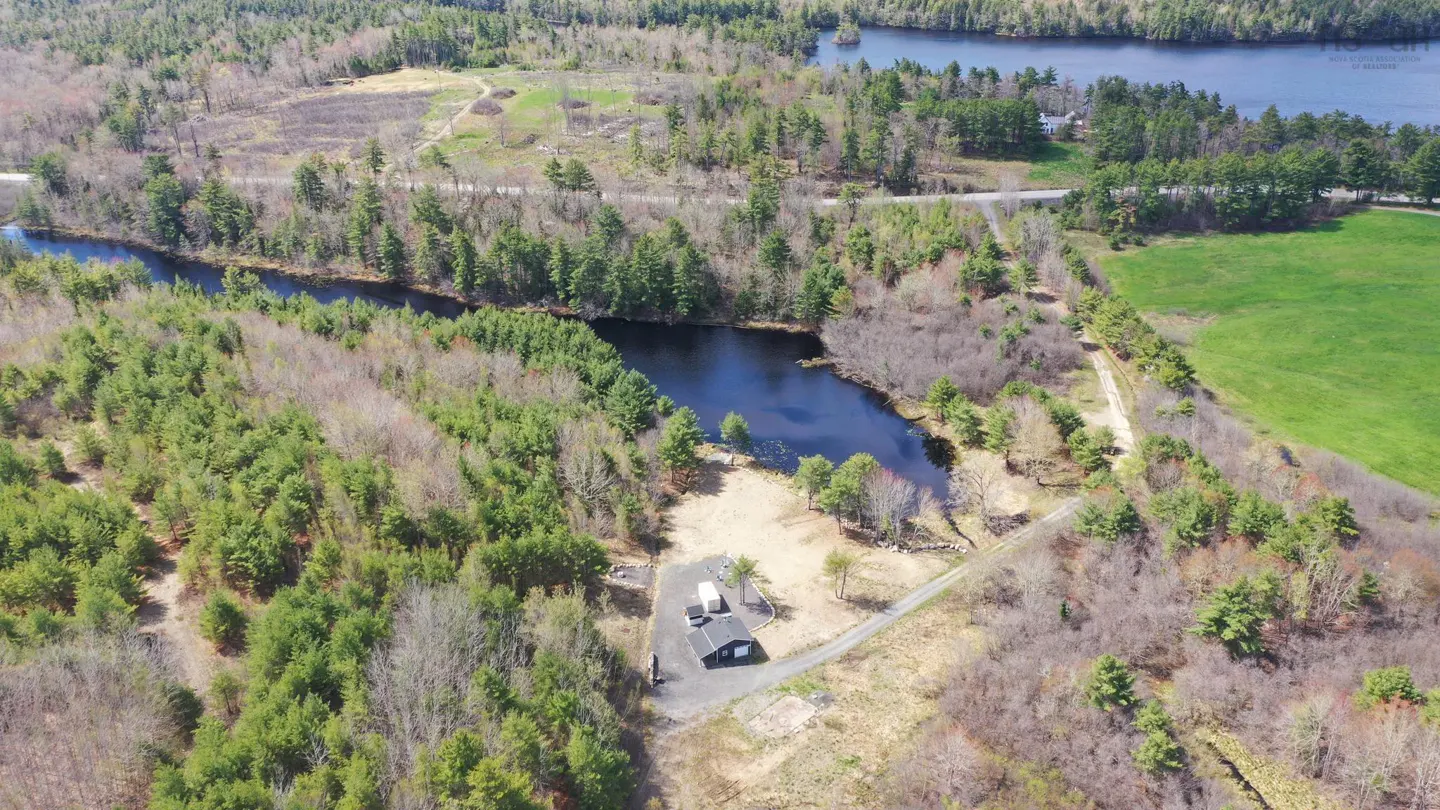 Aerial view of a dark-roofed house near a river, surrounded by green trees and brown fields. A lake is visible in the background.