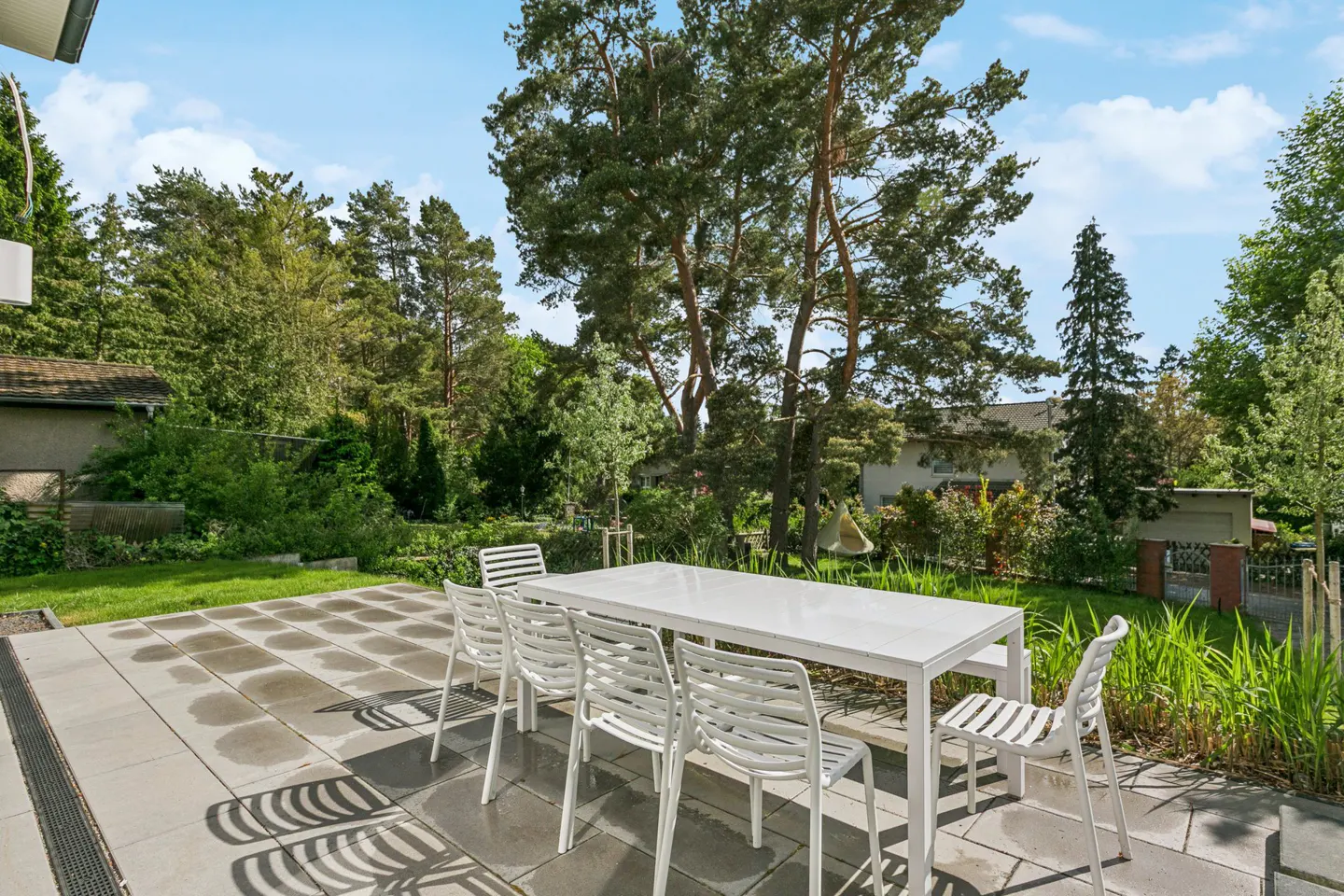 Outdoor patio with a white table and chairs on a stone patio, surrounded by green grass and trees.