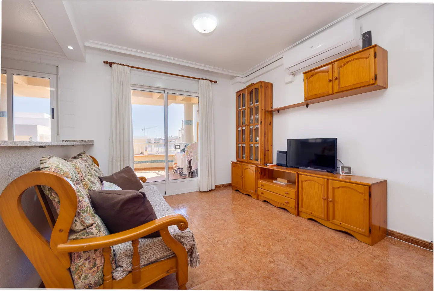 A living room with a wooden sofa, pillows, and a view of a balcony through a sliding glass door. A wooden cabinet and TV stand are on the right.