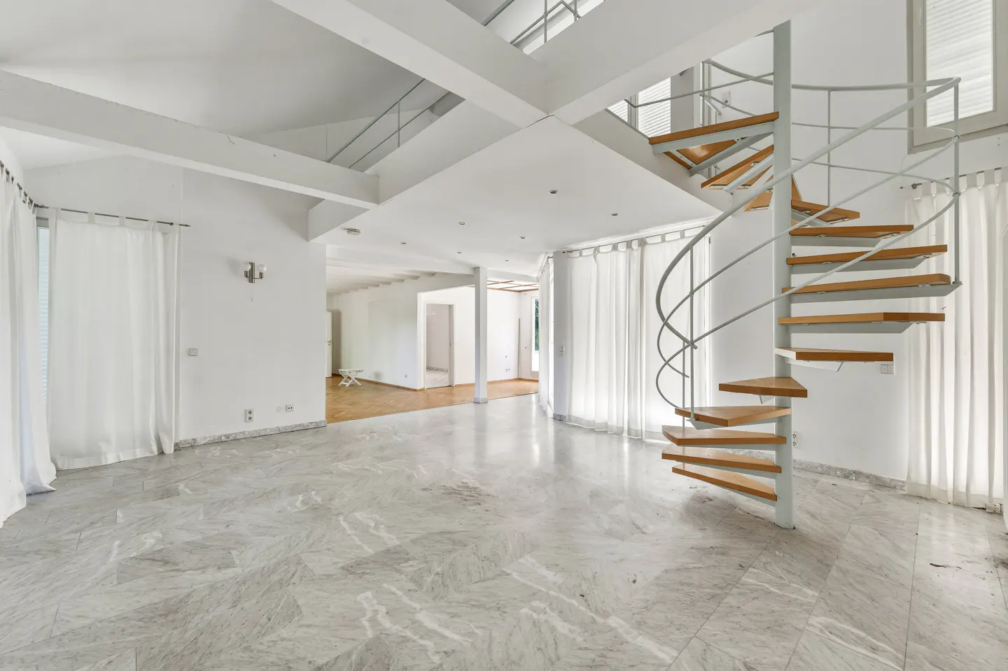Bright, airy room with marble floors, white walls, and a spiral staircase with wooden steps. White curtains hang by the windows.