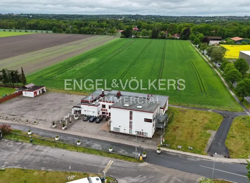 Aerial view of a white commercial building with a parking lot, adjacent to green fields under a cloudy sky.
