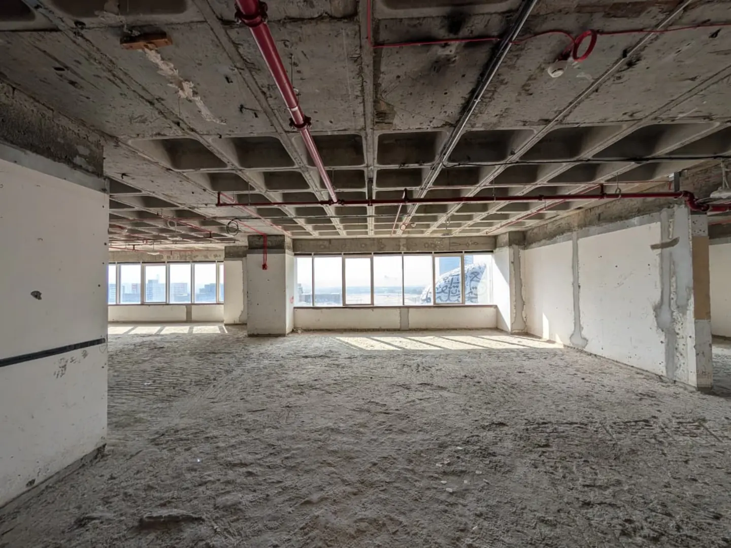 Unfinished office space with concrete floors, walls, and ceiling. Large windows offer a city view. Red pipes run along the ceiling.