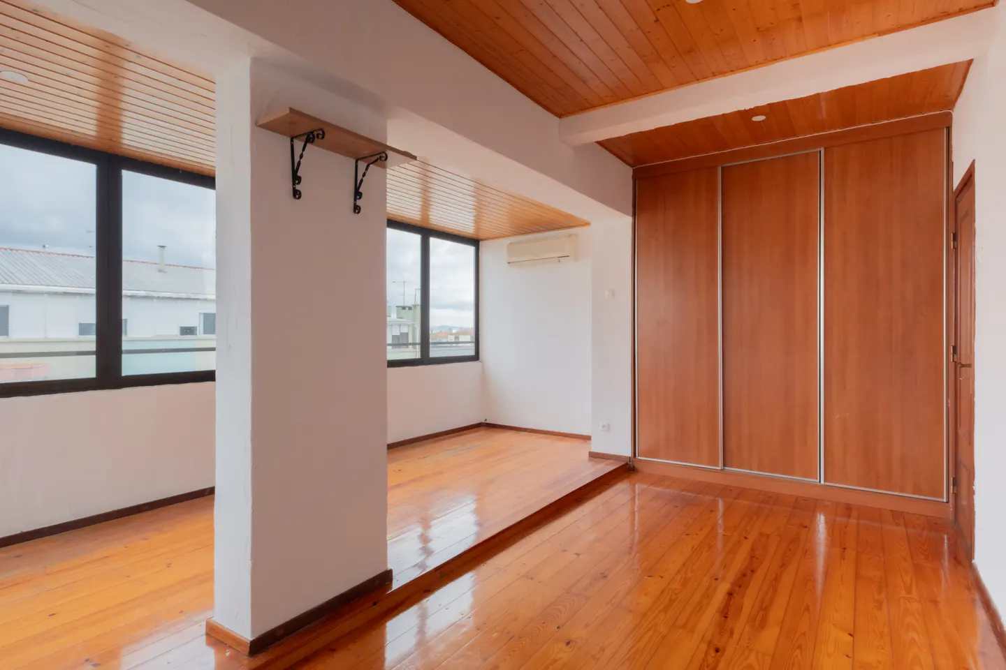 Bright room with wood floors, ceiling, and large closet. Windows offer a city view. A shelf is mounted on a white pillar.