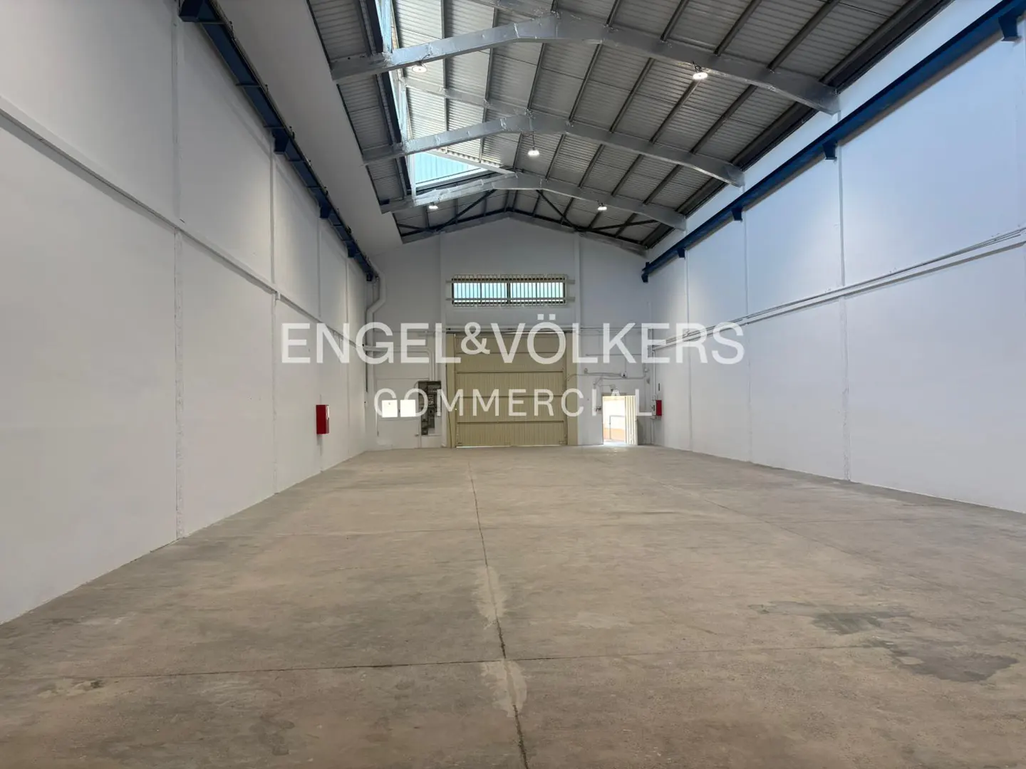 Empty, spacious industrial warehouse with white walls, concrete floors, and a high metal-truss ceiling with skylights.