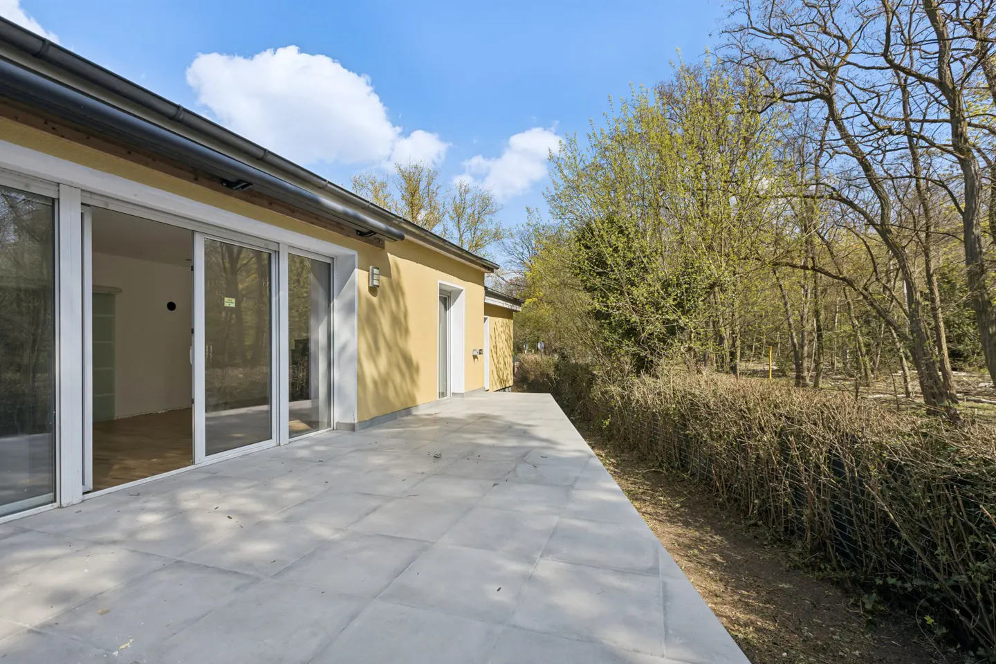 Exterior view of a yellow house with a gray tiled patio and sliding glass doors, backing onto a wooded area under a blue sky.