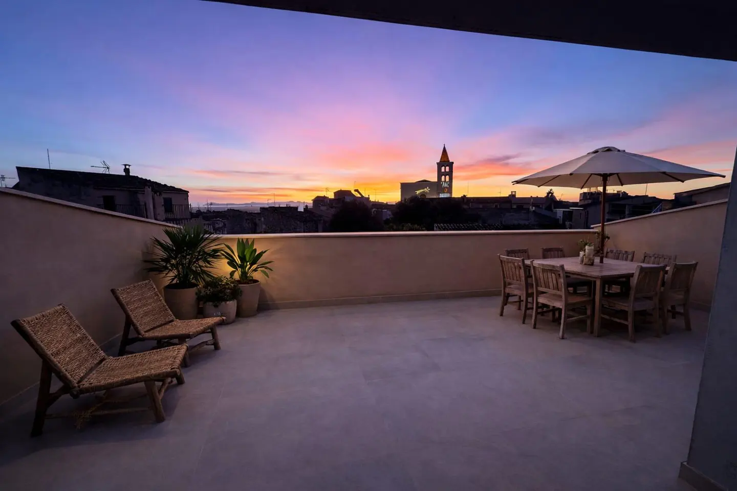 Rooftop patio with wicker chairs, dining table under an umbrella, and potted plants. A sunset sky with a church steeple is in the background.