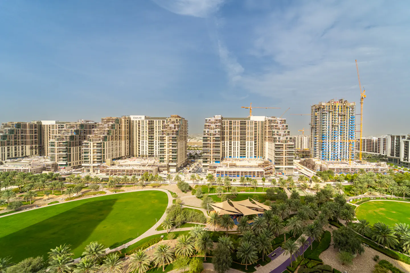 View of modern apartment buildings surrounding a green park with palm trees under a blue sky.