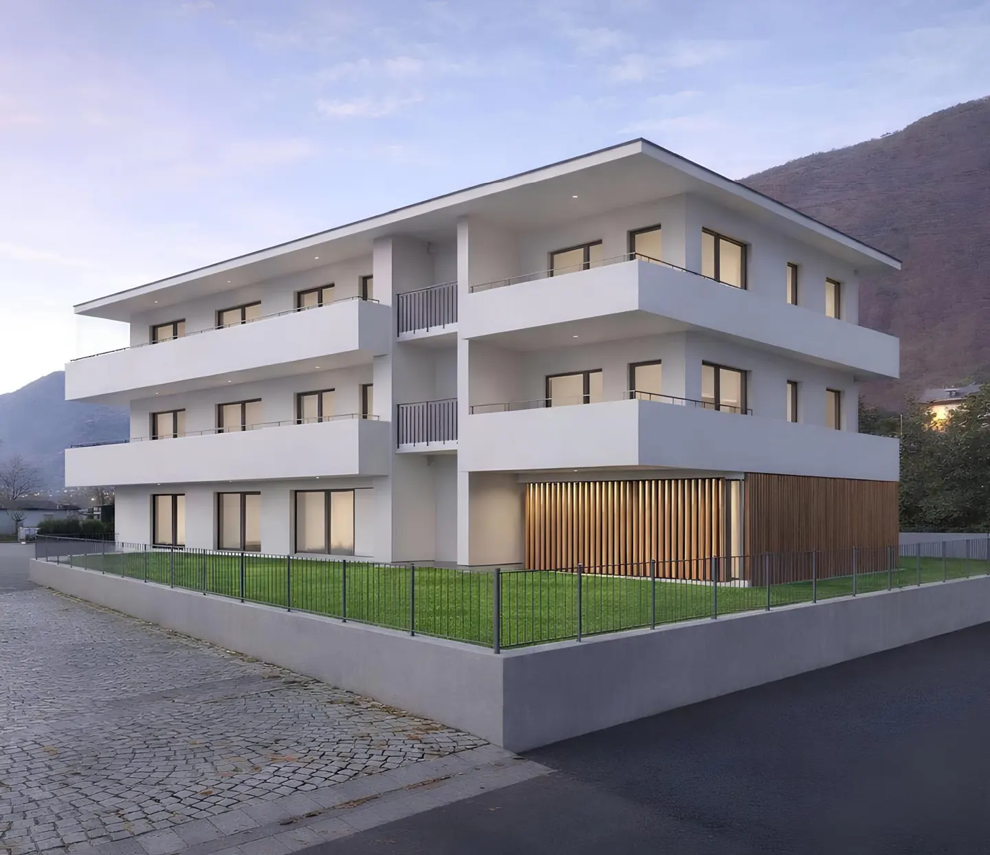 Modern three-story white apartment building with balconies, green lawn, and a mountain backdrop.