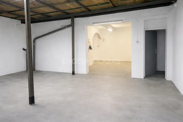 A bright, empty room with white walls and a gray concrete floor. A metal support beam is on the left. Another room is visible through an archway.
