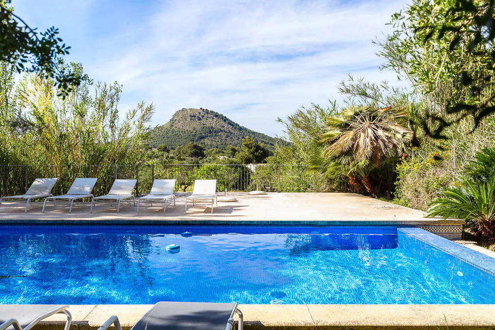 A bright blue tiled pool is surrounded by lounge chairs and lush greenery, with a mountain in the distance.