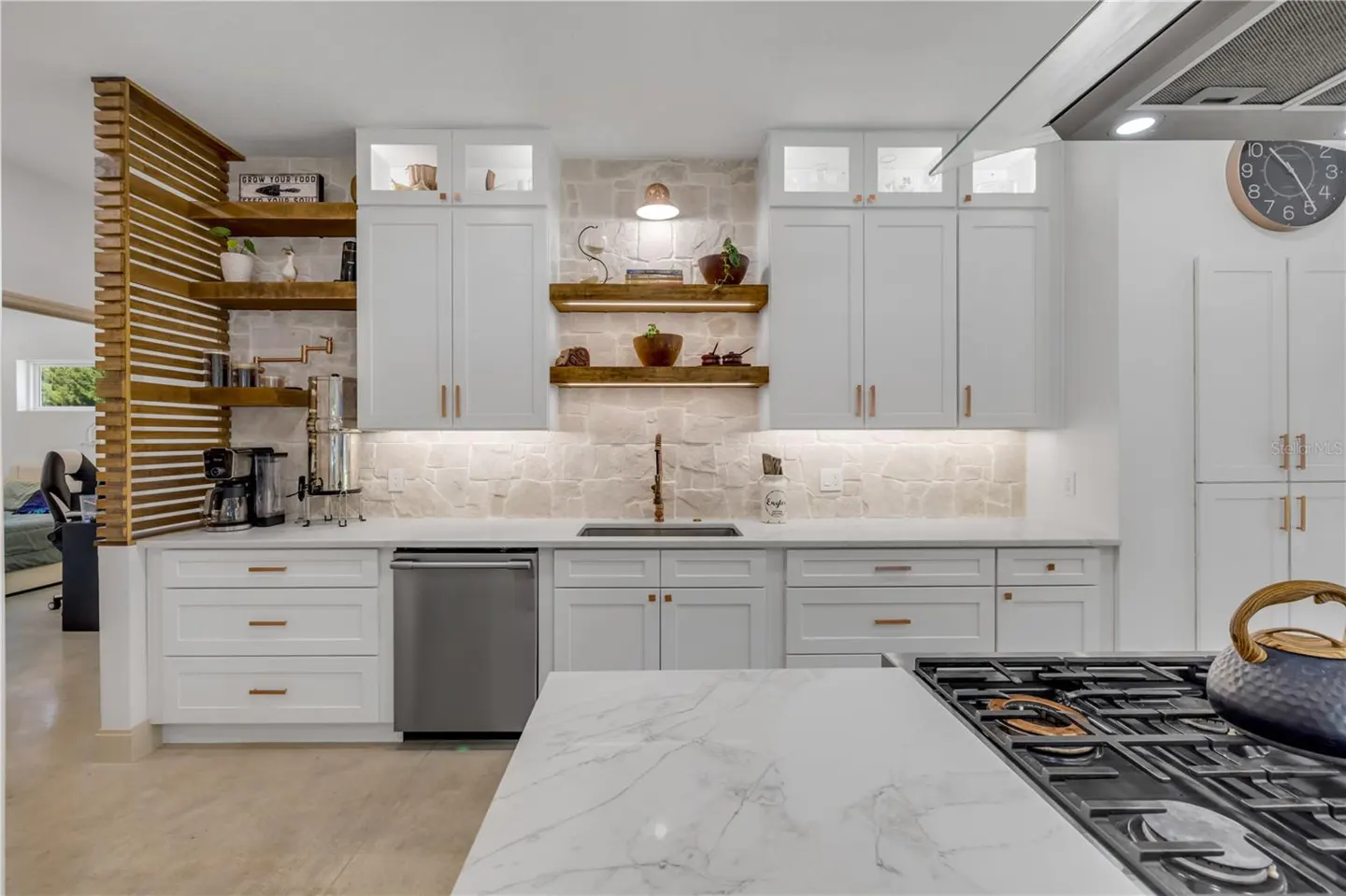 Bright kitchen with white cabinets, marble countertops, and wood shelves. A gas stove and kettle sit on the island.