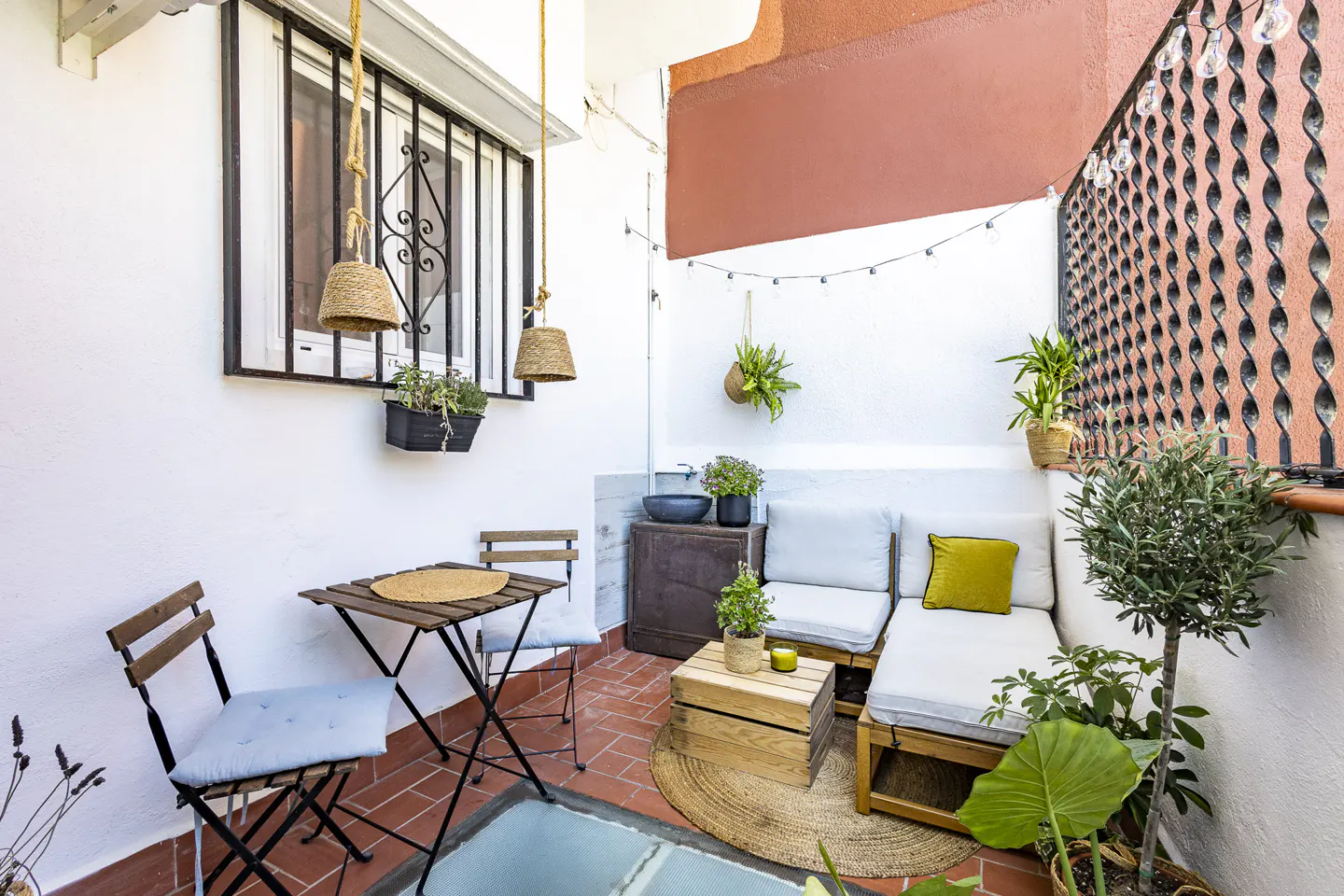 Cozy balcony with terracotta tiles, a small bistro set, wooden crate seating with white cushions, and hanging plants.