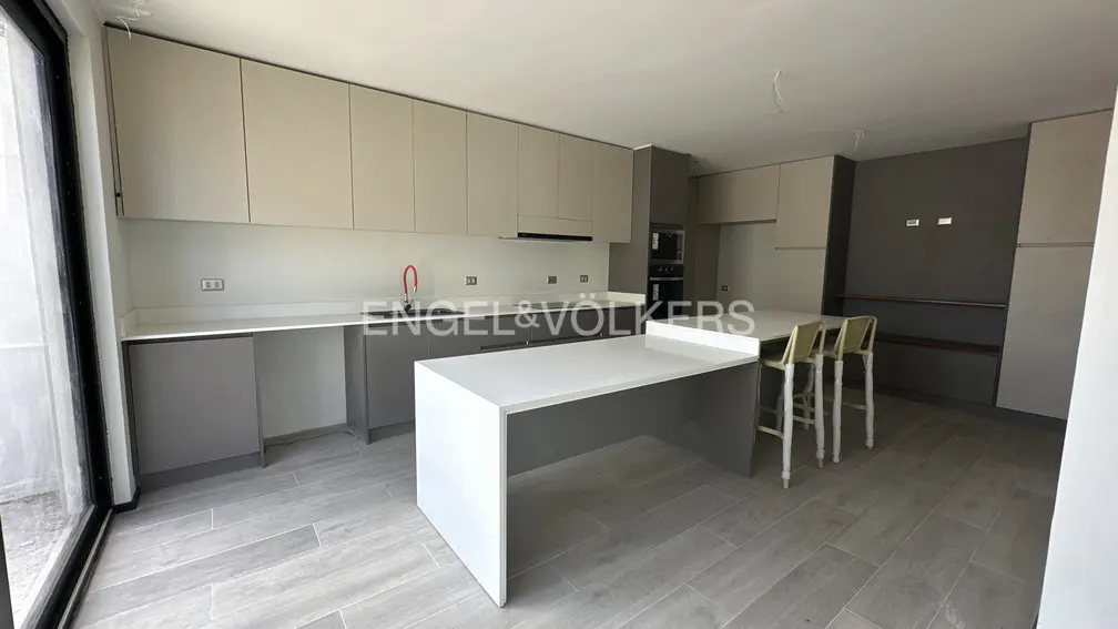 A modern kitchen with gray cabinets, white countertops, and a large island with seating. The floor is tiled in a light gray color.
