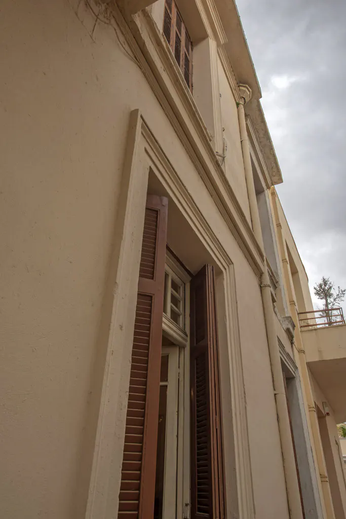 Low angle view of a beige building with brown shutters, some open, against a cloudy sky. Balcony with a tree is visible.